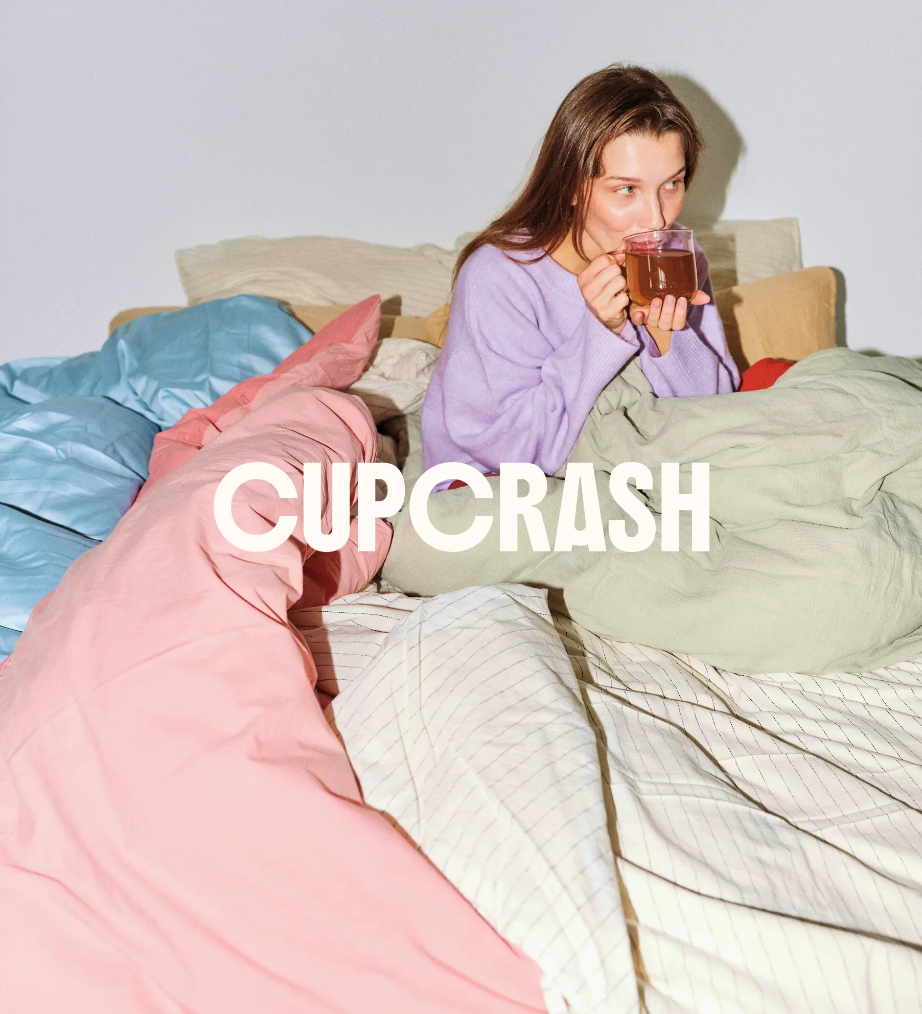 woman sitting among pastel-colored duvets, sipping tea peacefully - Cupcrash logo layered on top